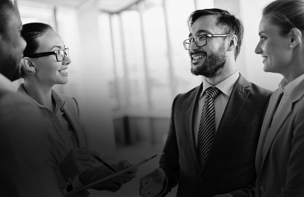 Black and white image of four people all smiling at one another dressed in smart clothing. One woman holds a pen and a clipboard.