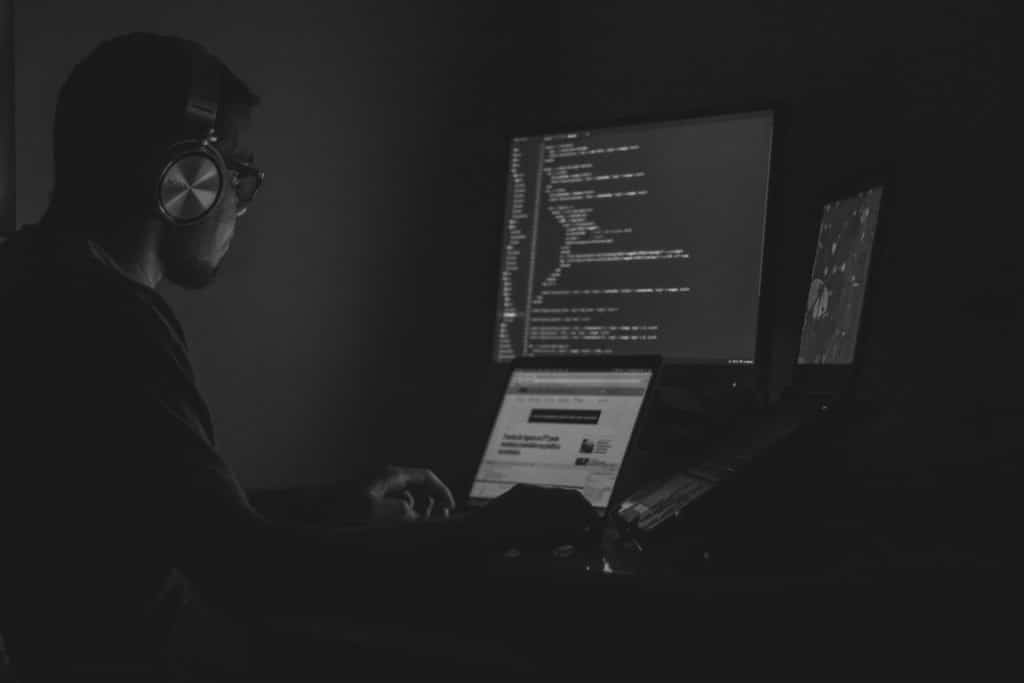 Black and white image of a man looking at a computer set up of three screens with lines of code on one of them. He wears headphones.