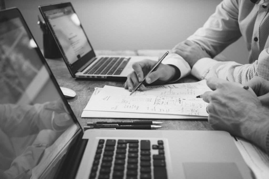 Black and white image of two people at a table with a pen and computer each. They are looking at a written on piece of paper.