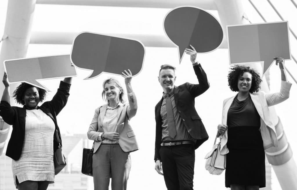 Black and white image of four people each wearing smart clothing and holding up speech bubble signs