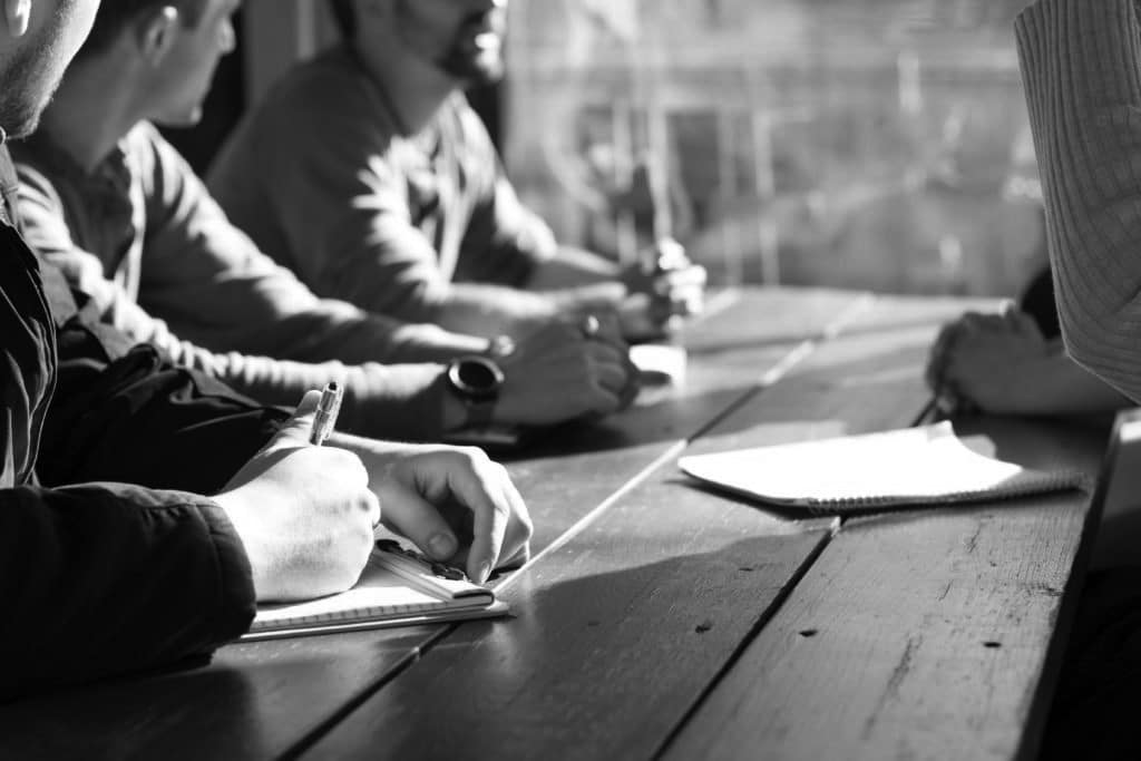 Black and white image of several people sat at a table with notepads and pens and their heads are turned to look at someone speaking